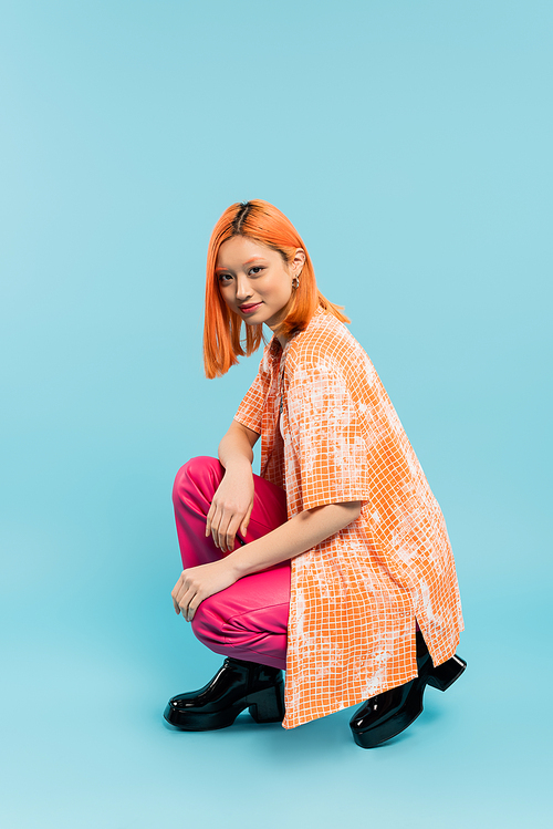 positive emotion, young asian woman with radiant smile looking at camera while posing on haunches on blue background, casual attire, orange shirt, pink pants, full length, generation z
