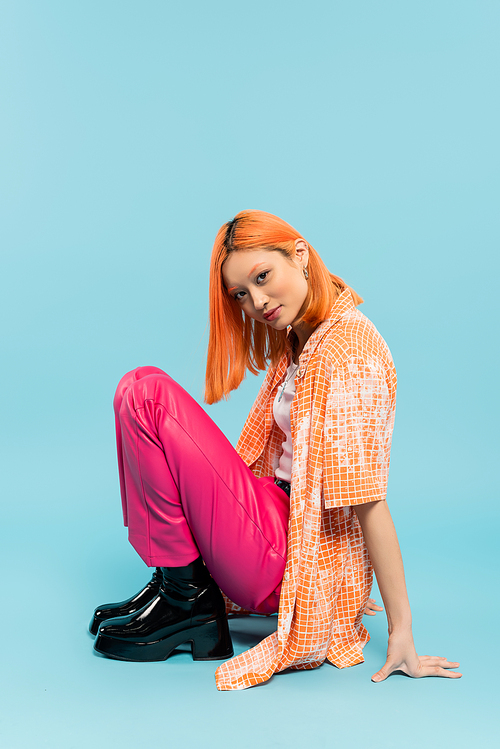 pleased face, summer vibes, youthful and fashionable asian woman with colored red hair looking at camera on blue background, casual clothes, orange shirt, pink pants, generation z