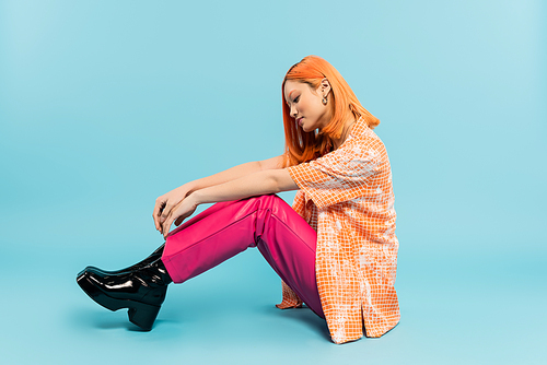 side view of young and stylish asian woman in orange shirt and pink pants sitting on blue background, dyed red hair, summer fashion, youthful style, full length, generation z
