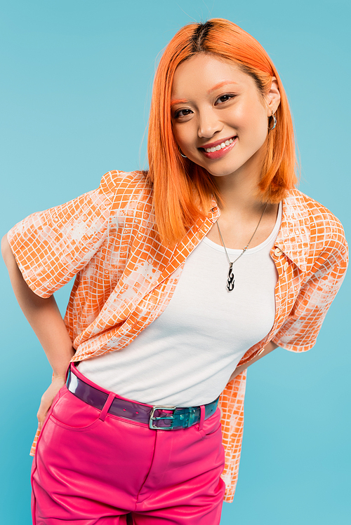 joyful and fashionable asian woman with colored red hair and radiant smile posing in orange shirt and looking at camera on blue background, happy summer, generation z lifestyle
