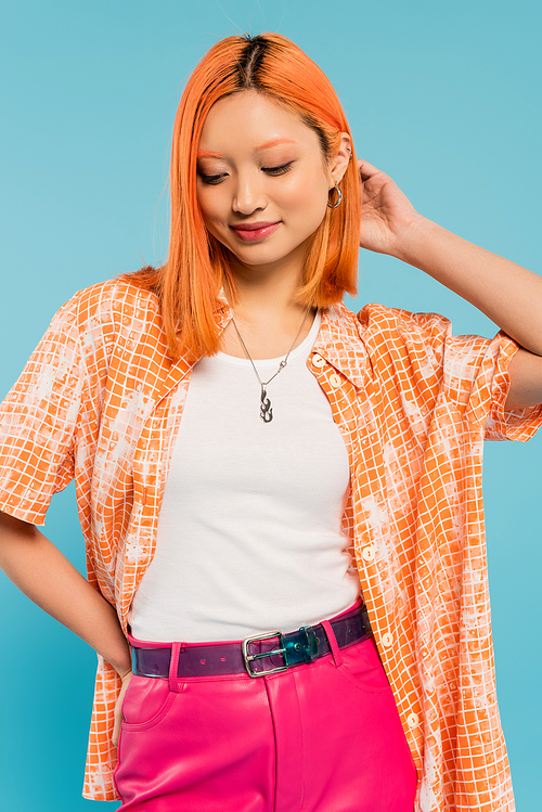 young and pleased asian woman with red colored red hair posing with hand on hip on blue background, orange shirt, pink pants, youthful fashion, summertime style, generation z