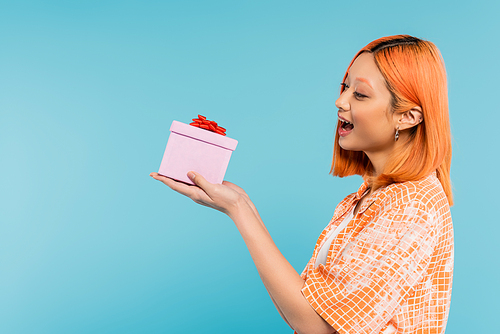 excitement and happiness, delighted young asian woman with colored red hair and open mouth holding festive present on blue background, stylish orange shirt, youthful fashion, side view