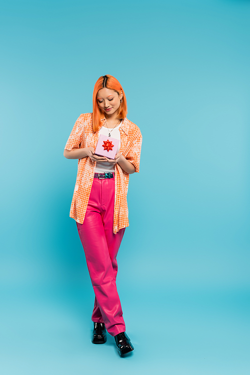 full length of young and joyful asian woman with happy smile, in trendy casual clothes standing with gift box on blue background, dyed red hair, orange shirt, pink pants, modern fashion