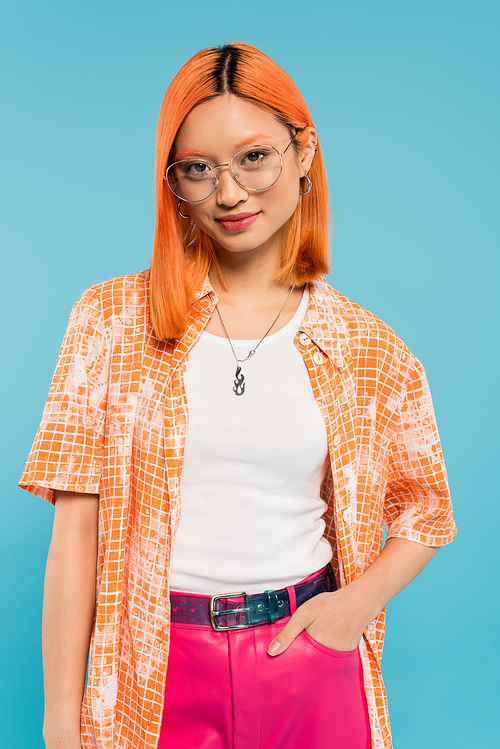 happy and trendy asian woman with colored red hair, in fashionable eyeglasses, orange shirt and pink pants standing with hand in pocket while smiling at camera on blue background