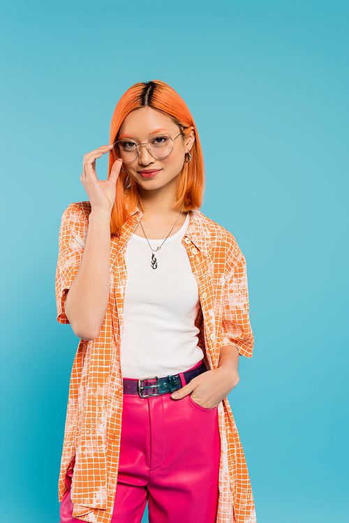 positivity, vibrant individuality, pleased asian woman adjusting trendy eyeglasses, holding hand in pocket and looking at camera on blue background, colored red hair, orange shirt, pink pants