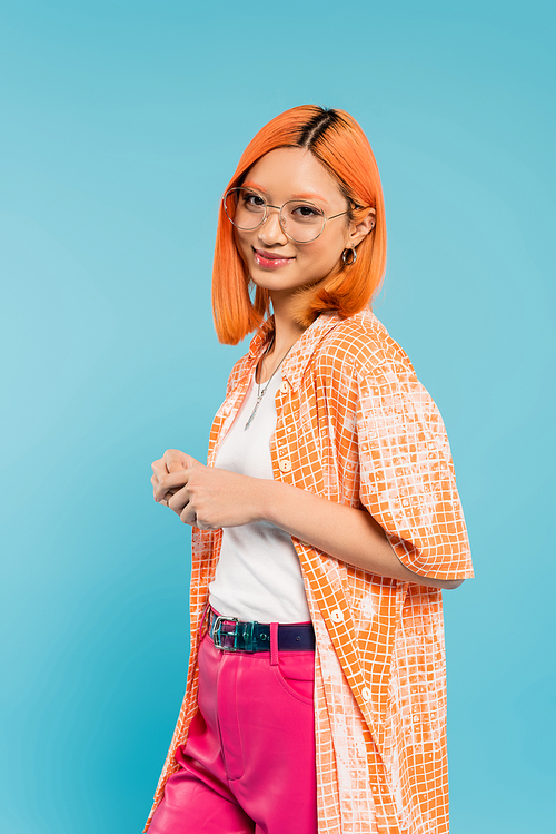 happy and fashionable summer, young asian woman with pleased face looking at camera while standing on blue background, dyed red hair, stylish spectacles, orange shirt, generation z lifestyle