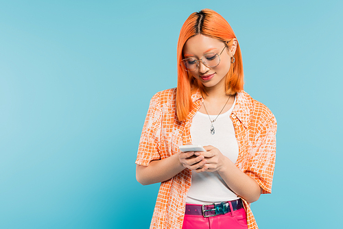 digital lifestyle, positive emotion, smiling asian woman with dyed red hair, in stylish eyeglasses and orange shirt networking on mobile phone on blue background, generation z, summer vibes