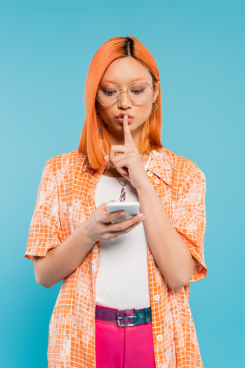 secrecy, confidentiality, serious asian woman in eyeglasses showing hush sign during video call on smartphone on blue background, red colored hair, orange shirt, modern fashion, generation z