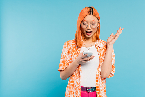 amazement, young asian woman with colored red hair and open mouth looking at mobile phone and gesturing on blue background, trendy eyeglasses, orange shirt, youthful fashion, digital lifestyle