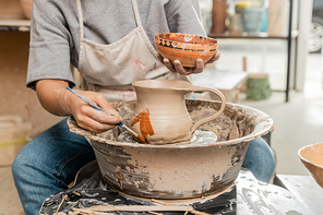 Cropped view of young female artist in apron painting on clay jug on pottery wheel near wooden tools n table in blurred ceramic workshop, clay shaping technique and process