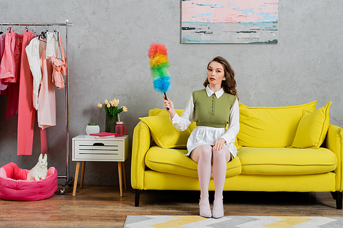 housekeeping concept, young woman with wavy hair sitting on couch and holding dust brush, housewife in dress and white tights looking at camera, domestic life, posing like a doll