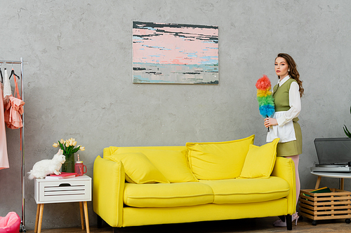 housekeeping concept, young woman with brunette wavy hair holding dust brush, housewife doing her daily duties, lifestyle, domestic chores, standing and looking at camera, painting on wall