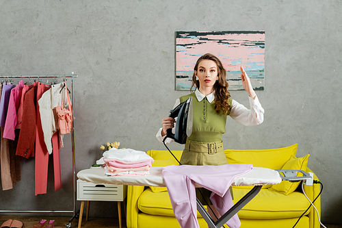 young woman with wavy hair acting like a doll, gesturing unnaturally, housewife holding iron near clean clothes and looking at camera, housekeeping concept, laundry day, daily routine