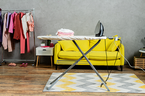 housekeeping concept, stack of folded and clean clothes near iron on ironing board, modern living room, domestic life, laundry day, yellow sofa, domestic chores