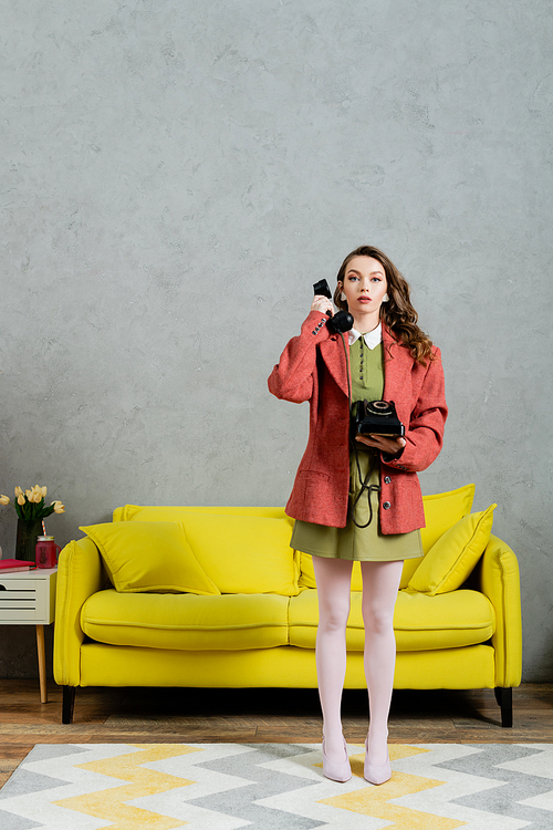 vintage vibes, beautiful woman with brunette and wavy hair standing near yellow couch, housewife talking on retro telephone, posing like a doll, looking at camera, modern interior, living room