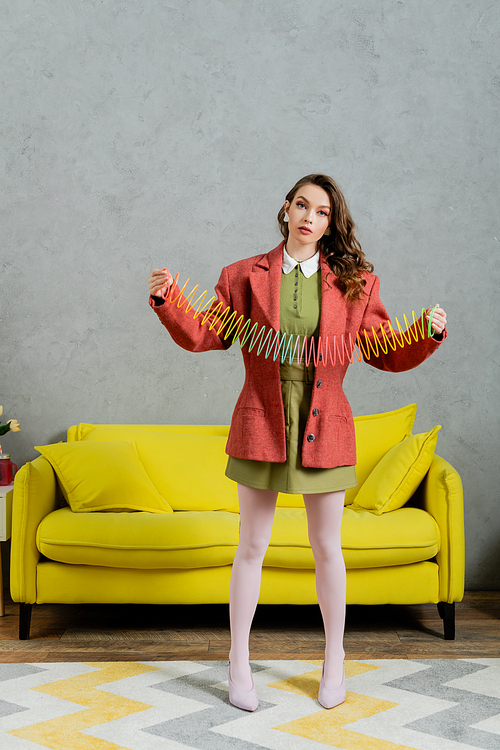 young brunette woman posing like a doll and playing with rainbow slinky, looking at camera, modern living room with yellow couch, childish, vintage, nostalgia, colorful toy, leisure and fun