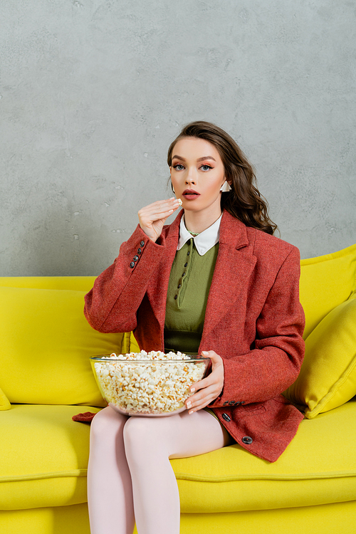 pretty girl acting like a doll, concept photography, young woman with brunette wavy hair eating popcorn, holding bowl, salty snack, home entertainment, sitting on comfortable yellow sofa