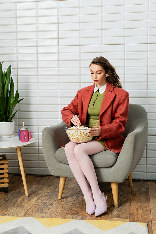 concept photography, young woman with brunette wavy hair sitting on comfortable armchair, acting like a doll, looking at bowl with popcorn, leisure, home entertainment, movie snack