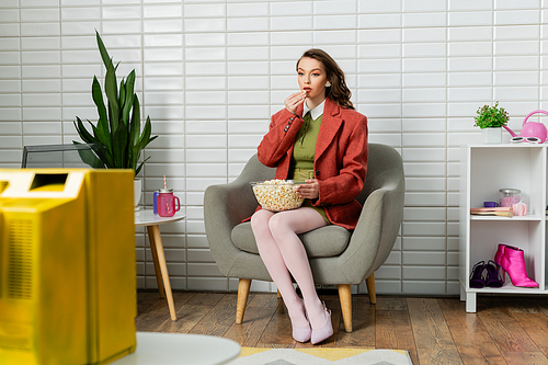 concept photography, young woman with brunette wavy hair sitting on comfortable armchair, watching tv and eating popcorn, leisure, home entertainment, movie snack, doll like