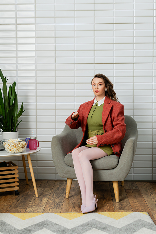 concept photography, young woman with brunette wavy hair sitting on comfortable armchair next to bowl with popcorn, acting like a doll and gesturing unnaturally, movie snack, looking at camera