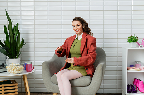 concept photography, happy young woman with wavy hair sitting on comfortable armchair next to bowl with popcorn, acting like a doll and gesturing unnaturally, movie snack, looking at camera