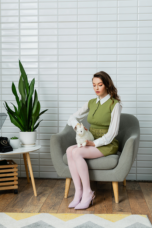 woman acting like a doll, beautiful woman sitting on comfortable grey armchair and holding toy rabbit, green plants and retro telephone on table, concept photography