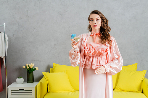 concept photography, woman acting like a doll, domestic life, housewife in pink outfit with silk robe holding cocktail in glass, gesturing and standing near yellow coach in modern living room