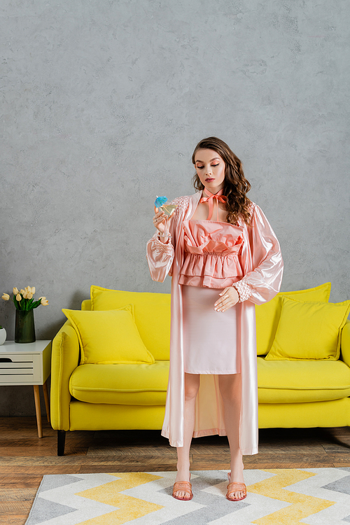 concept photography, young woman acting like a doll, domestic life, housewife in pink outfit with silk robe holding cocktail in glass, gesturing and standing near yellow coach in modern living room