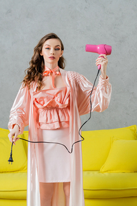 concept photography, woman acting like a doll, beautiful housewife in pink silk robe holding hair dryer and plug, standing near yellow coach in modern living room, pretending like drying hair