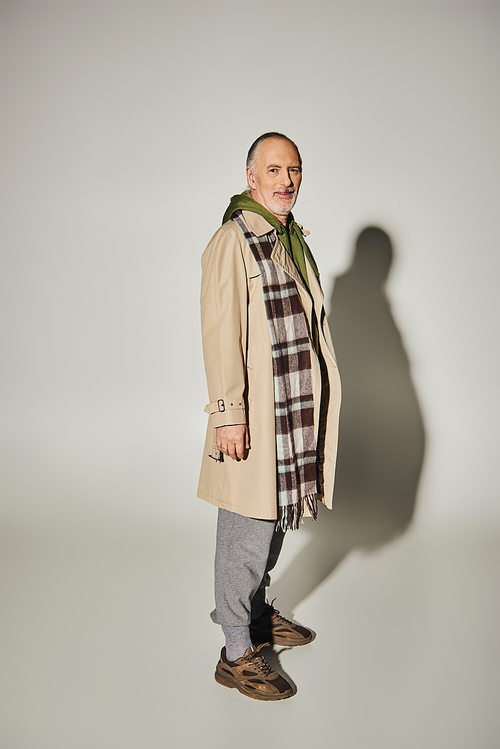 full length of glad senior model with grey hair and beard standing in trendy casual wear and smiling at camera on grey background with shadow, beige trench coat, plaid scarf, happy aging concept