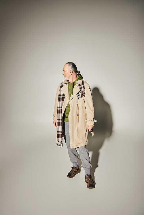 full length of aged and bearded man in beige stylish trench coat, green hoodie and plaid scarf standing and looking away on grey background with shadow and copy space, fashion and age concept
