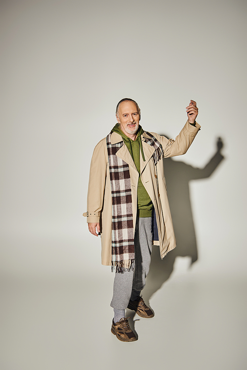 full length of expressive and optimistic senior man in green hoodie, plaid scarf and beige trench coat gesturing and looking at camera on grey background with shadow, happy age concept