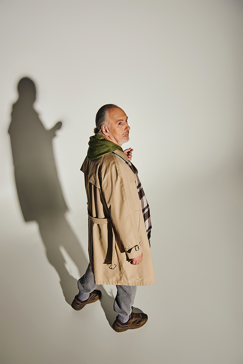 full length of thoughtful senior man in green hoodie, beige trench coat and plaid blazer looking away on grey background with shadow, fashionable lifestyle, high angle view