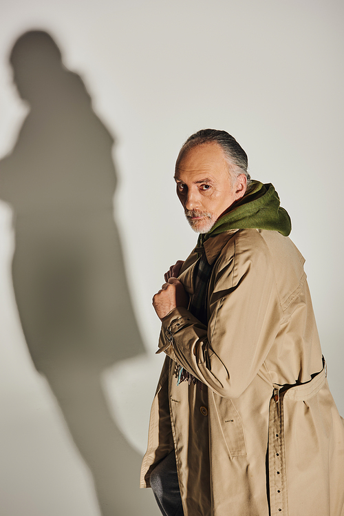 fashion and age, personal style, confident senior man with serious face expression standing in beige trench coat and looking at camera on grey background with shadow
