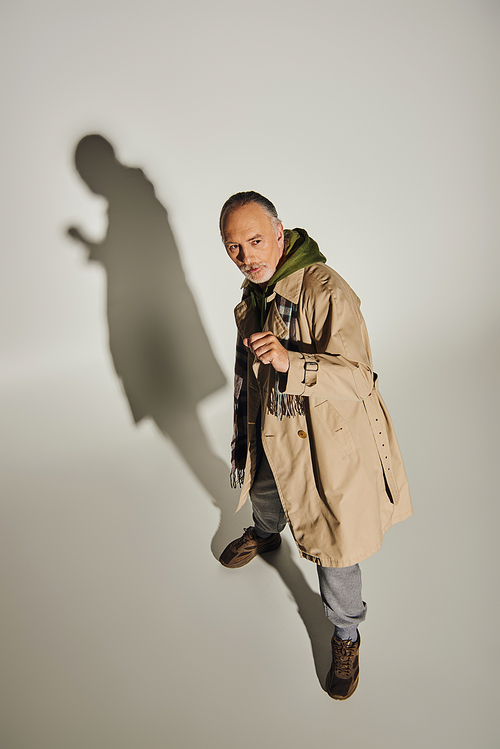high angle view of expressive and stylish senior male model looking away on grey background with shadow, grey haired man in beige trench coat, plaid scarf and green hoodie, fashion and age concept