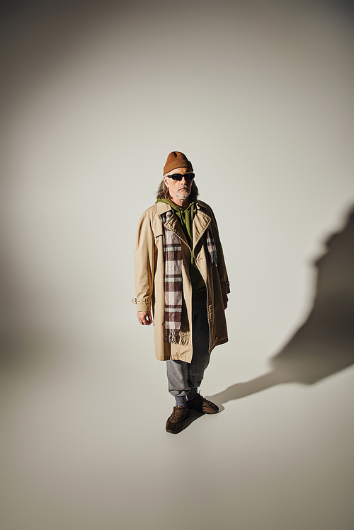 fashion and age concept, full length of hipster style senior man in beanie hat, dark sunglasses, beige trench coat and plaid scarf standing on grey background with shadow and copy space