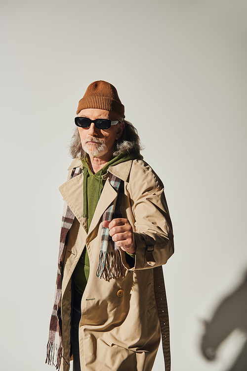 confident senior man in dark sunglasses and hipster style attire standing with clenched fist and looking at camera on grey background, beanie hat, beige trench coat, fashionable aging concept