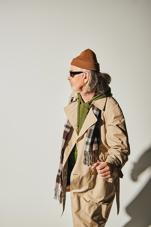 stylish senior male model standing and looking away on grey background, aged hipster man in dark sunglasses, beanie hat, beige trench coat and plaid scarf, fashionable lifestyle concept
