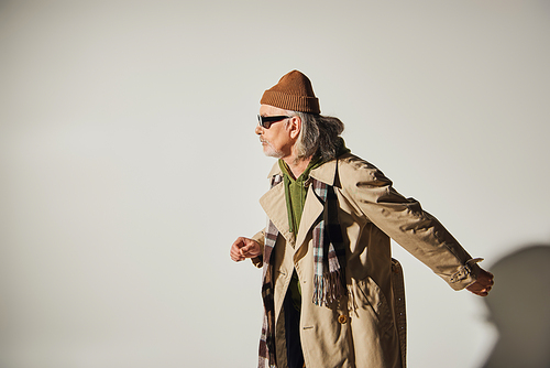side view of senior man in trendy casual clothes walking on grey background, beanie hat, dark sunglasses, beige trench coat, plaid scarf, hipster style, fashionable aging concept