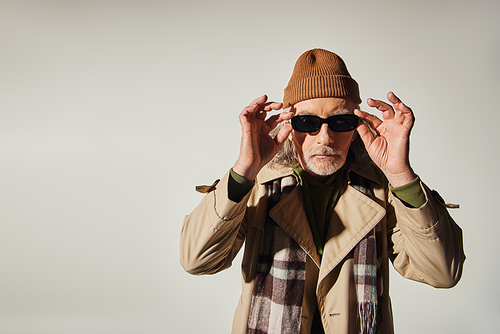 fashionable aging, hipster style, cool senior man in beanie hat, plaid scarf and trench coat adjusting dark sunglasses and looking at camera on grey background, fashion shoot