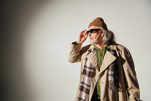 elderly man in beanie hat, beige trench coat and plaid scarf adjusting dark sunglasses and looking away on grey background, hipster style, individuality, fashion and age concept