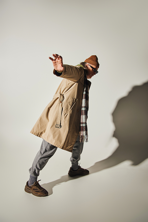 full length of senior hipster man showing stop gesture while standing in expressive pose on grey background with shadow, dark sunglasses, beanie hat, beige trench coat, fashion and age concept