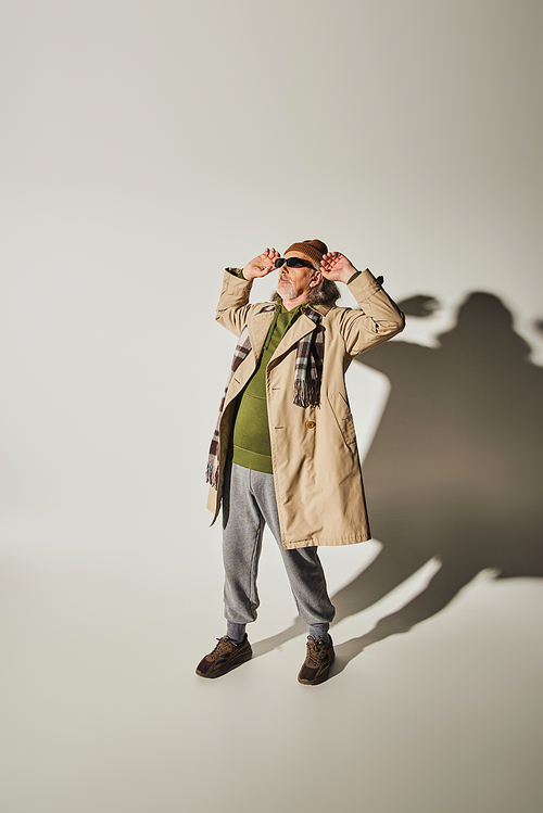 full length of cool senior man in beanie hat and beige trench coat adjusting dark sunglasses and looking away on grey background with shadow, casual hipster style attire, fashion and age concept