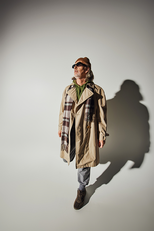 hipster style, fashionable aging concept, high angle view of senior man in beanie hat, dark sunglasses, beige trench coat and plaid scarf posing on grey background with shadow