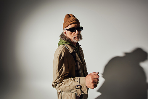 senior male model in dark sunglasses and stylish casual clothes looking at camera on grey background with shadow, beanie hat, beige trench coat, plaid scarf, expressive personality