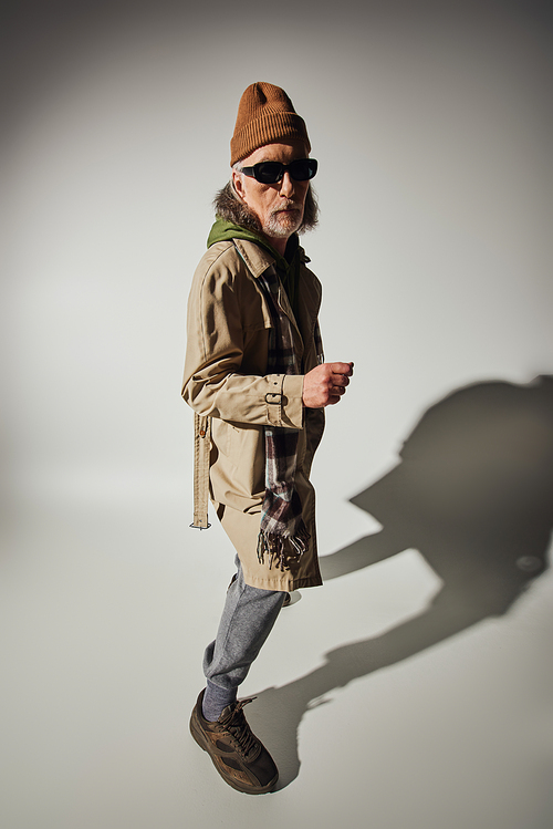 aging with style, senior hipster man in dark sunglasses, beanie hat, green hoodie and beige trench coat looking at camera on grey background with shadow, positive aging and fashion concept