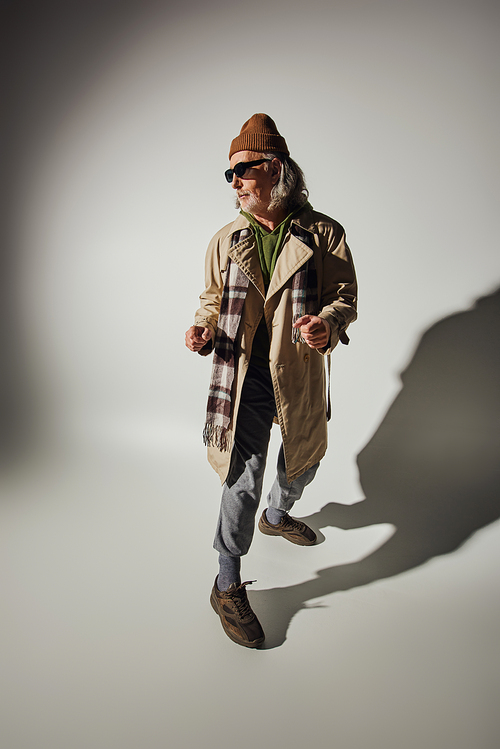 expressive personality, senior hipster man in dark sunglasses, beanie hat and beige trench coat standing in stylish pose on grey background with shadow, positive and trendy aging concept