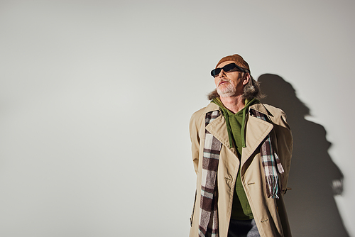 elderly man in stylish casual clothes and dark sunglasses standing on grey background with shadow and looking away, hipster trend, beanie hat, beige trench coat, fashion and age concept