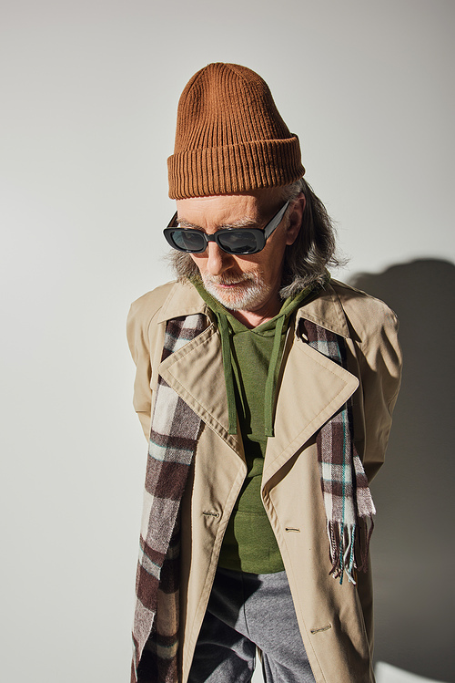 senior male model, hipster style clothes, beanie hat, dark sunglasses, plaid scarf, beige trench coat, individuality, fashion and age concept, grey background with shadow