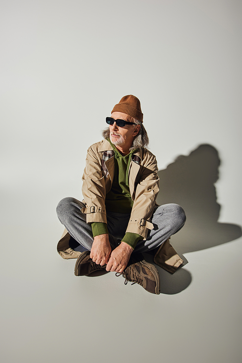full length of senior man in hipster style attire and dark sunglasses sitting with crossed legs on grey background with shadow, beanie hat, beige trench coat, sneakers, fashion and age concept